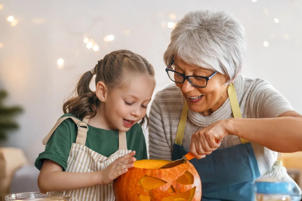 A young girl carves a pumpkin with an older lady, possibly her grandparent, looking on and helping with the knife.